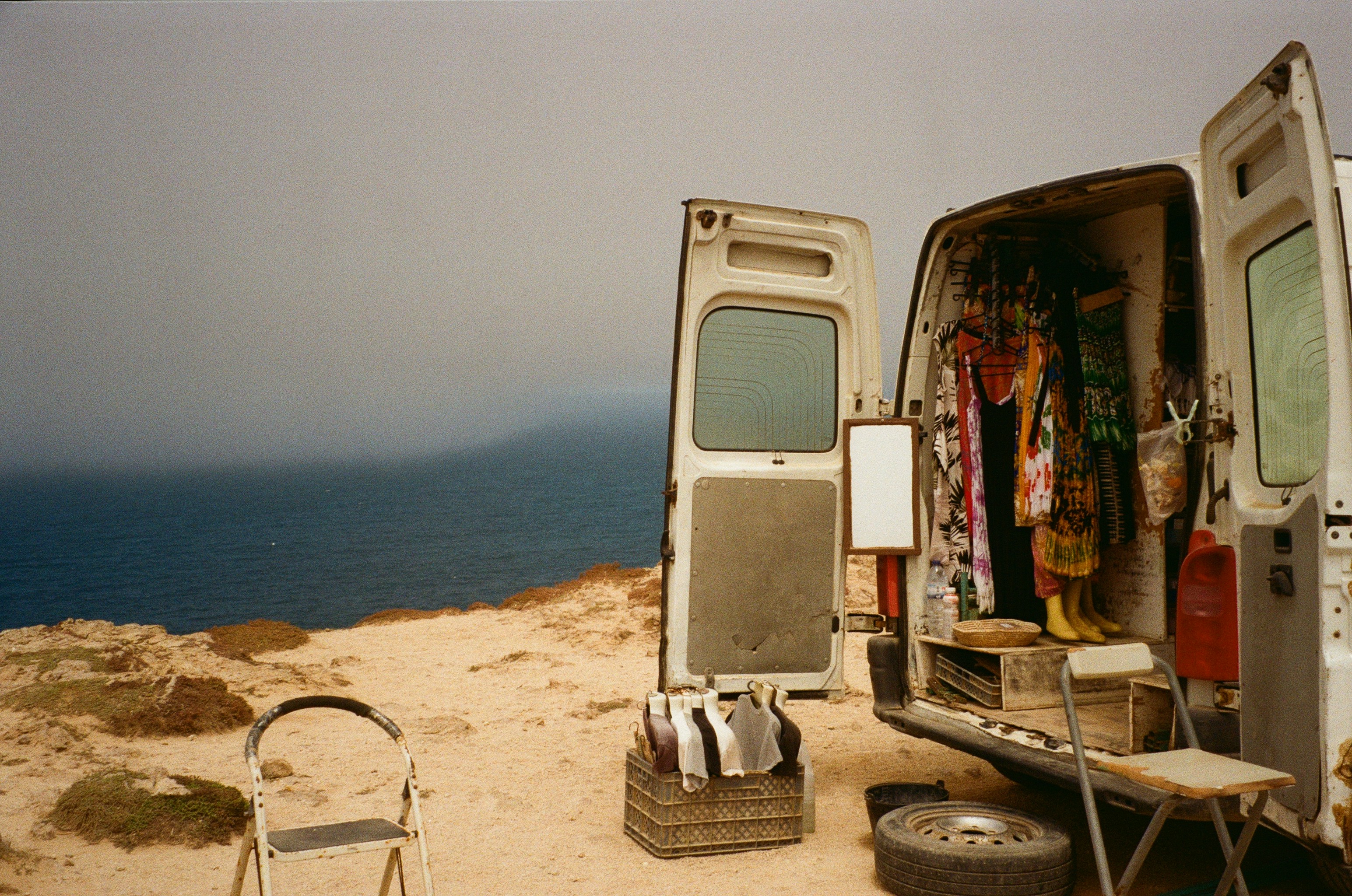 A vintage van with open doors reveals a rack of colorful clothes, parked on a cliff edge above the sea along the Lycian Coast